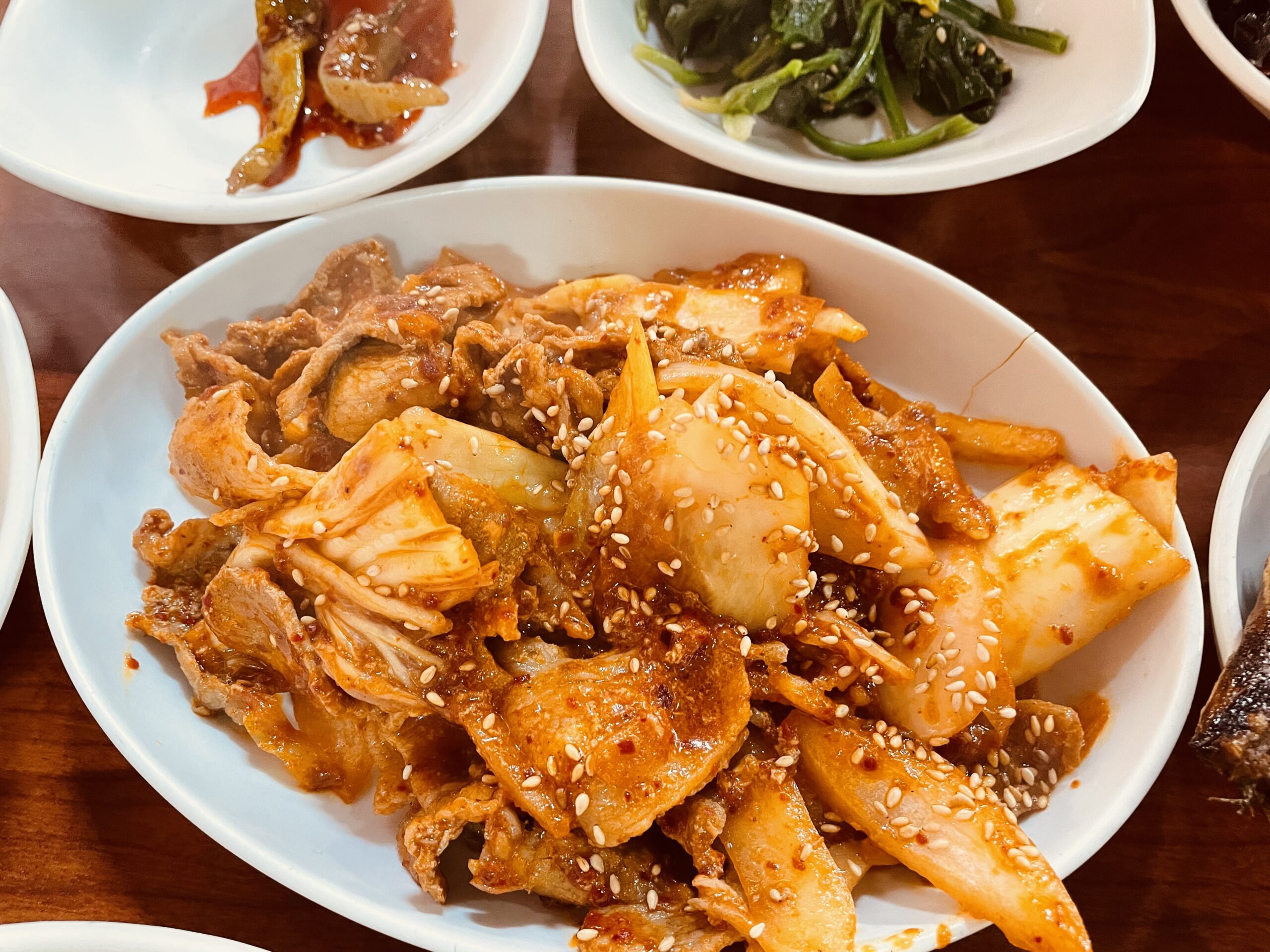 Spicy stir-fried pork with kimchi and onion, sprinkled with sesame seeds, served with Korean banchan side dishes